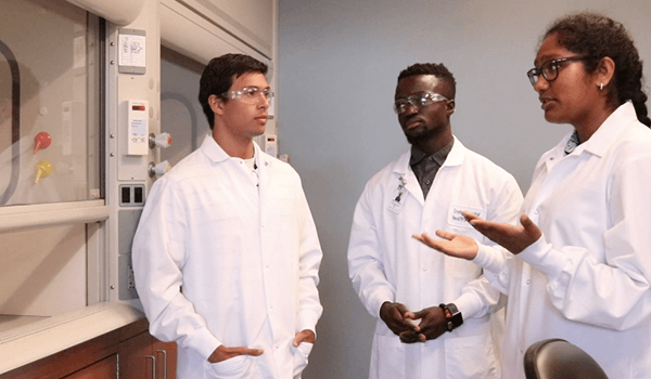 The image shows three people in white lab coats talking to each other in front of a fume hood sash