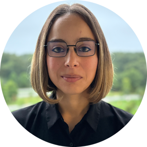 Headshot of Lina Marcella Carmona wearing glasses and a bob haircut, in a black shirt. Picture is set against a natural outdoor background