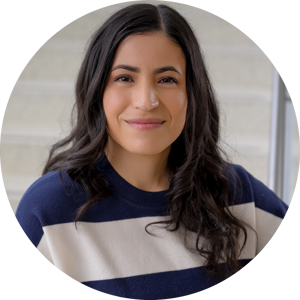 A headshot of Jennifer Vega with long dark hair in a navy and white striped sweater, smiling softly in front of a light-colored staircase
