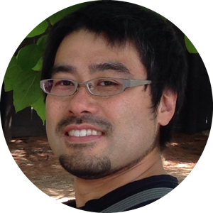 A selfie of Tadatoshi Sato wearing a broad smile in his glasses. He has a short dark hair and a goatee. The photo is set in front of large green leaves as a background.