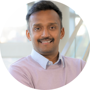 A headshot of Varanasi Siva Karthik, smiling broadly with arms crossed. He is wearing a lavender sweater over a light blue collared shirt, near a staircase with large glass windows in the background.