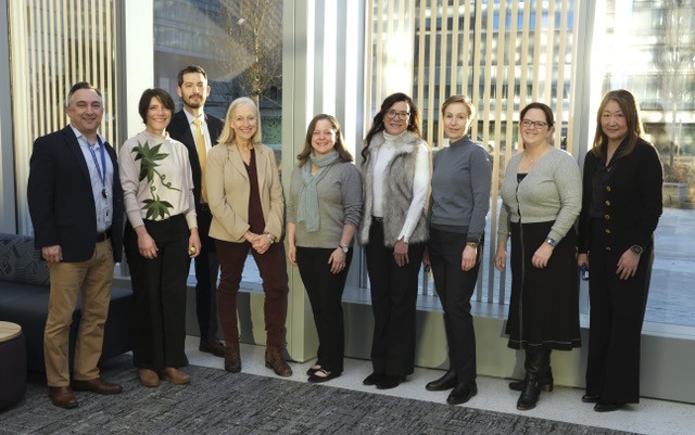 9 individuals standing in front of a large window smiling